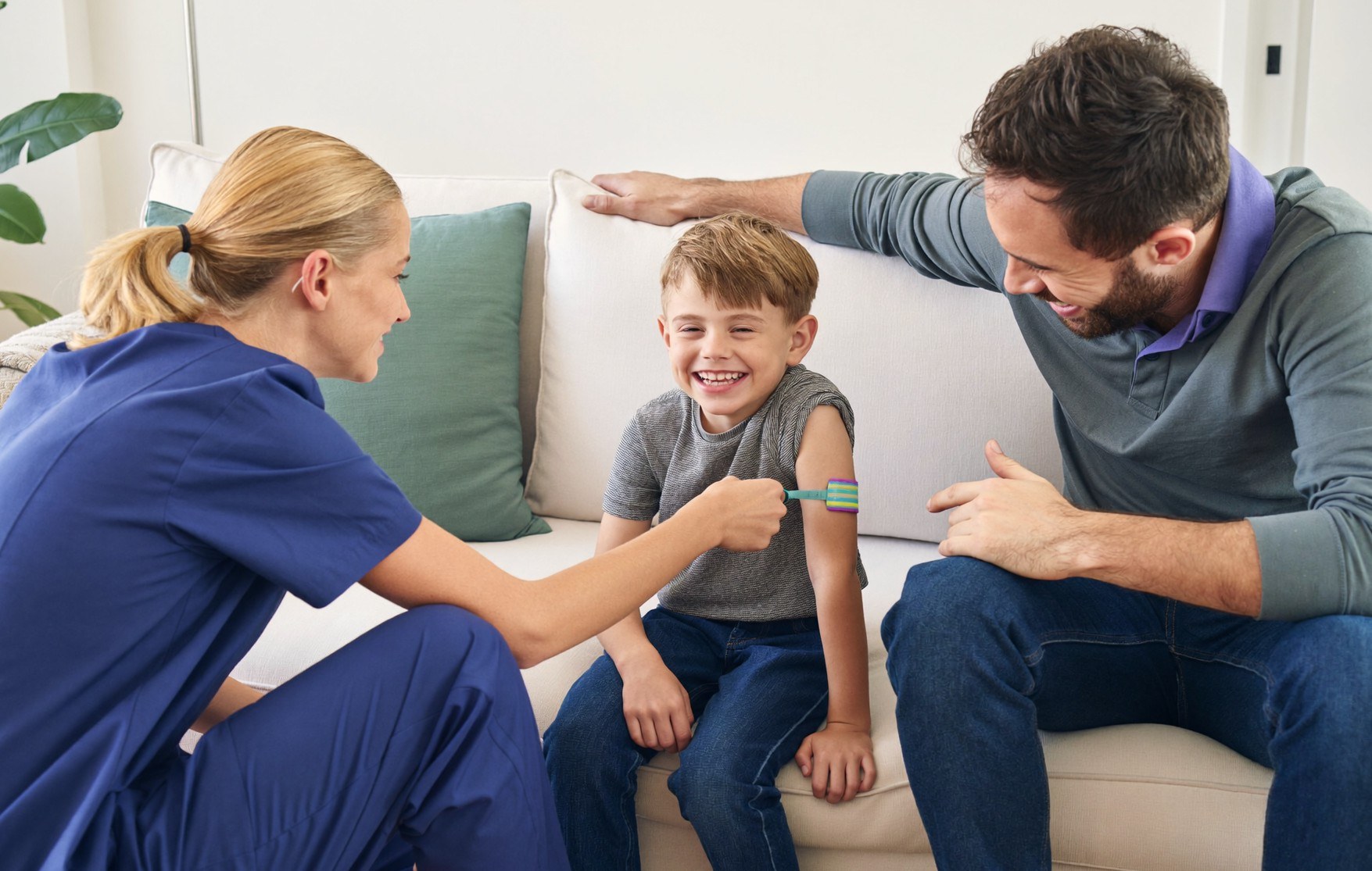 A smiling boy shows the nurse his bandage, his father beside him